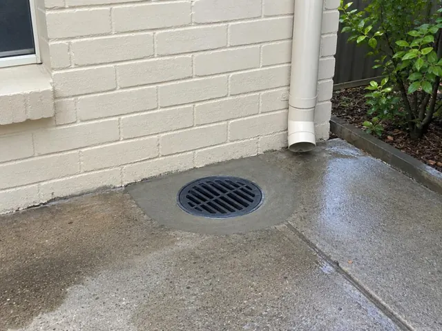 Outdoor drain grate beside a brick wall and downpipe on wet concrete in Mayfield East